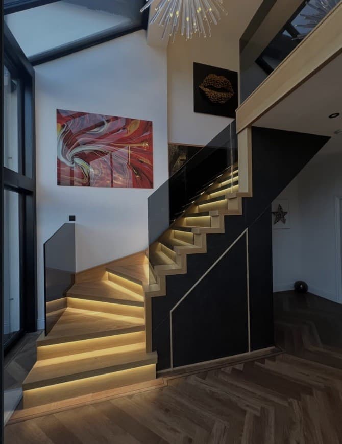 Bespoke oak staircase with warm integrated step lighting, frameless glass balustrades and dark under-stair panelling in a double-height entrance.