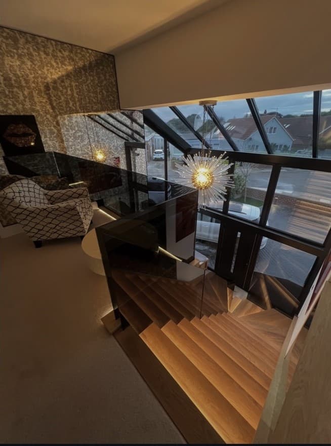 View down the illuminated oak staircase from the upper landing, with frameless glass and the glazed entrance visible below.