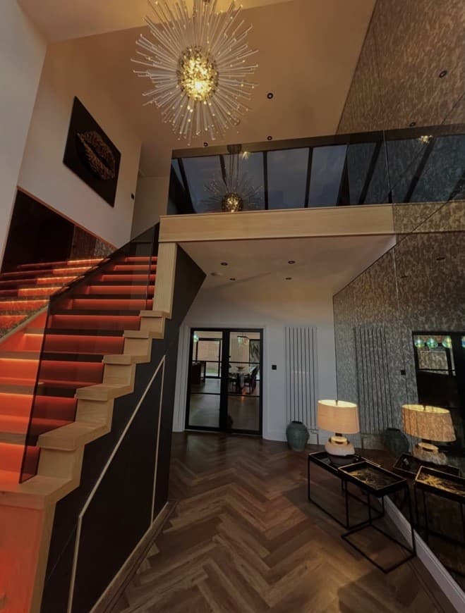 Double-height entrance hallway with illuminated staircase, glass balustrade and reflective feature wall.