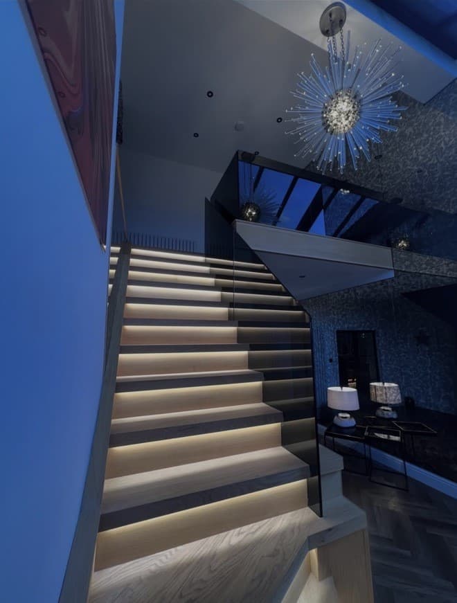 Upward view of the illuminated oak staircase and frameless glass balustrade beneath a statement pendant light.