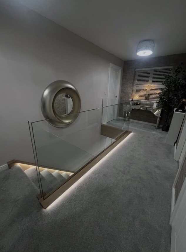 Carpeted upper landing with frameless glass stair balustrade and warm integrated lighting.