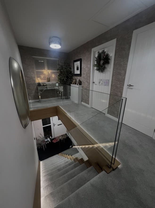 Upper landing view over the oak staircase with frameless glass balustrades and integrated perimeter lighting.