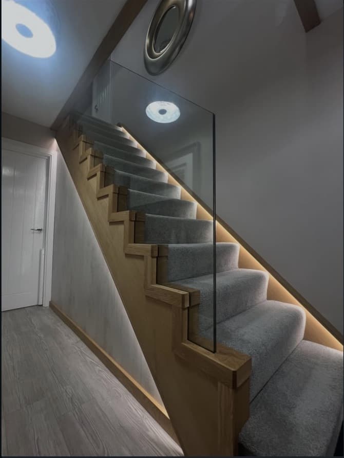 Angled view of a carpeted oak staircase with warm stringer lighting and a frameless glass balustrade.