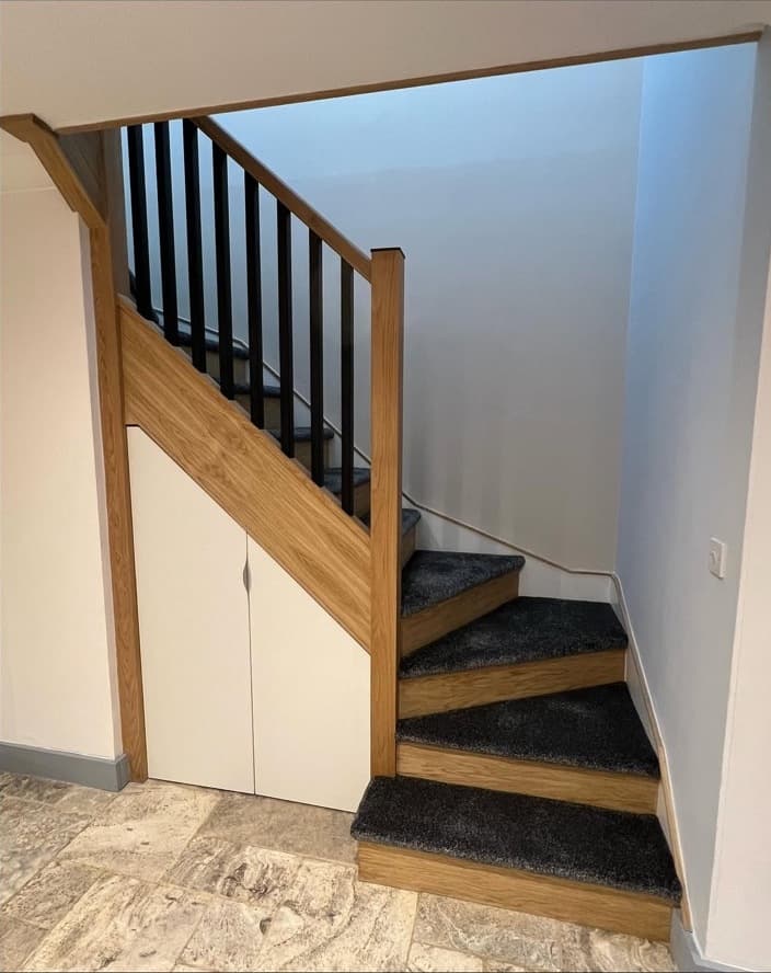 Finished oak staircase with black spindles, dark carpet and fitted under-stair storage.