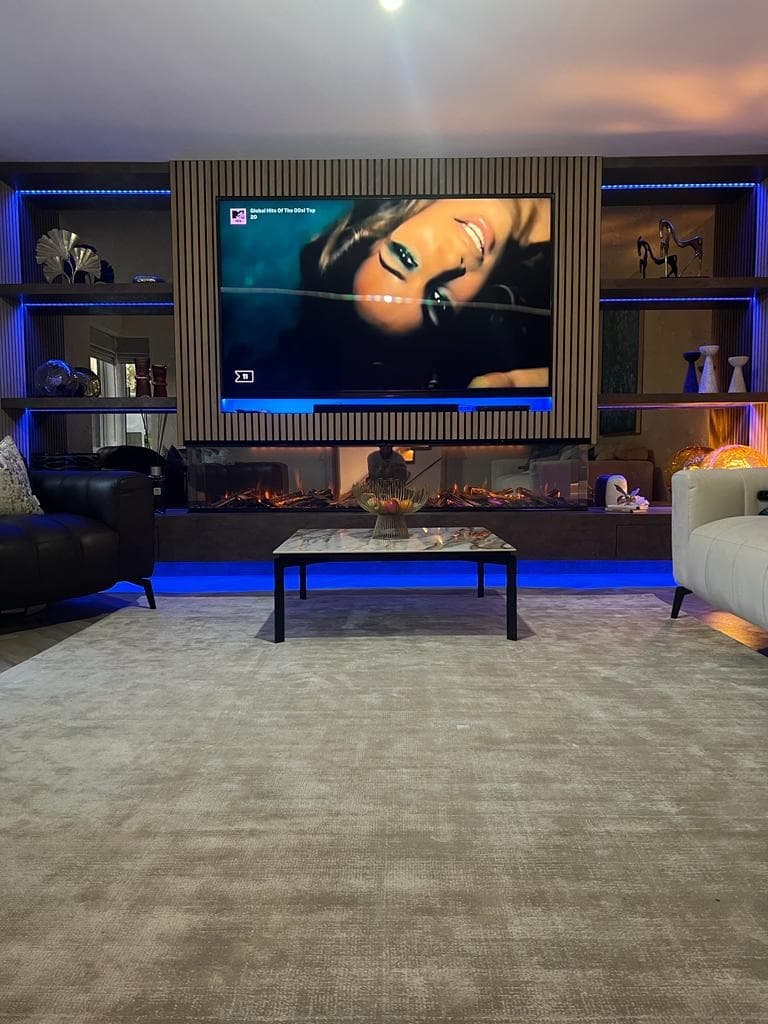 Front-on bespoke media wall with natural oak slatted acoustic panels, bronze mirror shelving, fireplace and blue LED lighting.