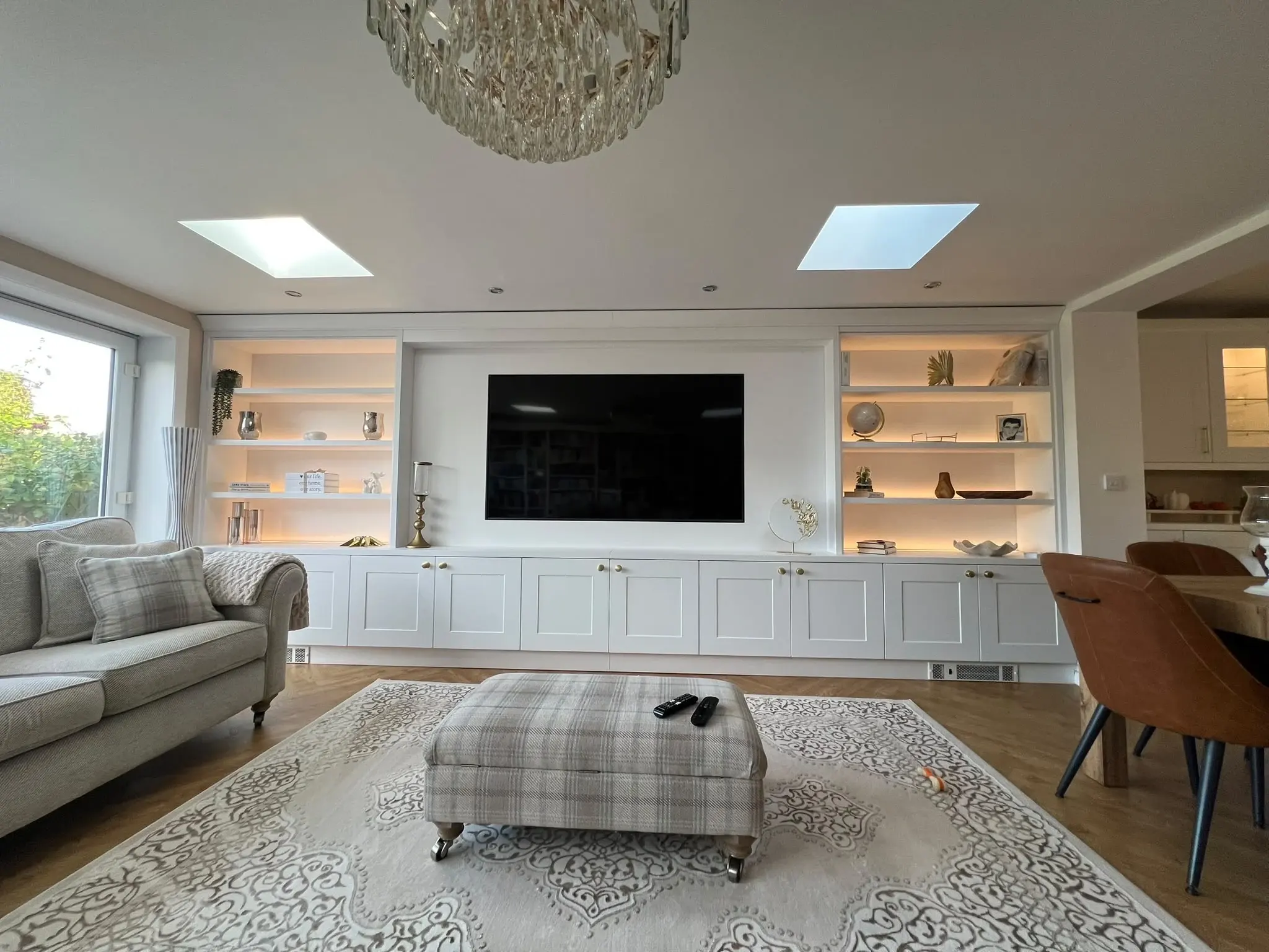 White fitted media wall with a television, warm illuminated shelves and a long run of lower cupboards.