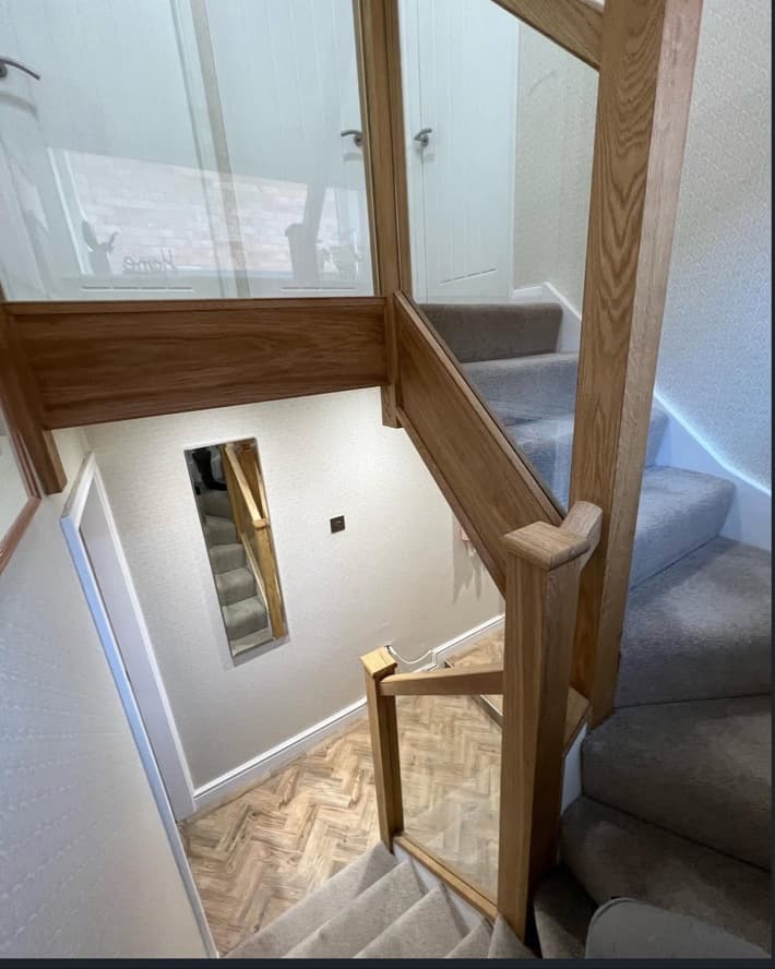 View down the renovated oak staircase with clear glass balustrades and grey carpet.